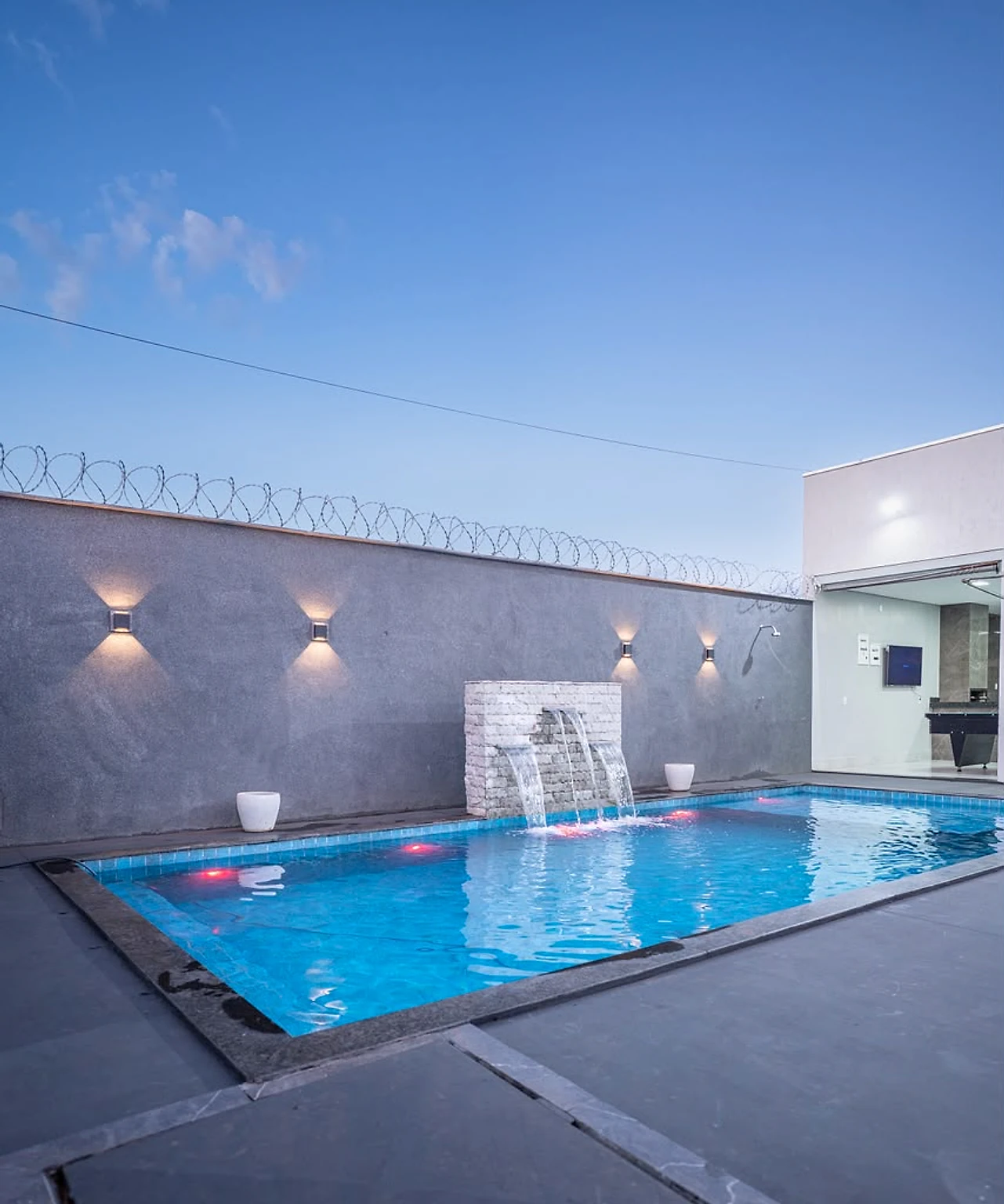 Piscina com cascata, luzes na água e paredes escuras ao entardecer
